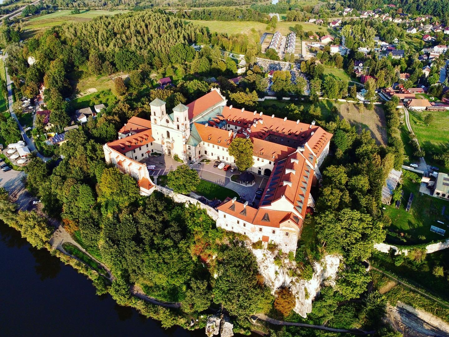 Opactwo Benedyktynów w Tyńcu z lotu ptaka, malowniczo położone na wapiennej skale nad Wisłą, otoczone zielenią drzew i zabudową mieszkalną w słoneczny dzień.