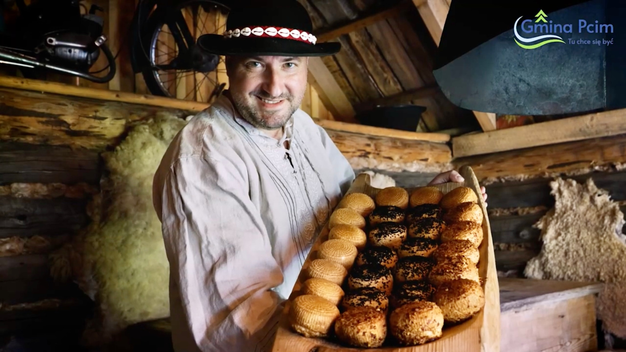 Uśmiechnięty mężczyzna w kapeluszu góralskim prezentuje tacę z oscypkami w drewnianej chacie. W tle widoczny rower i logo Gminy Pcim.