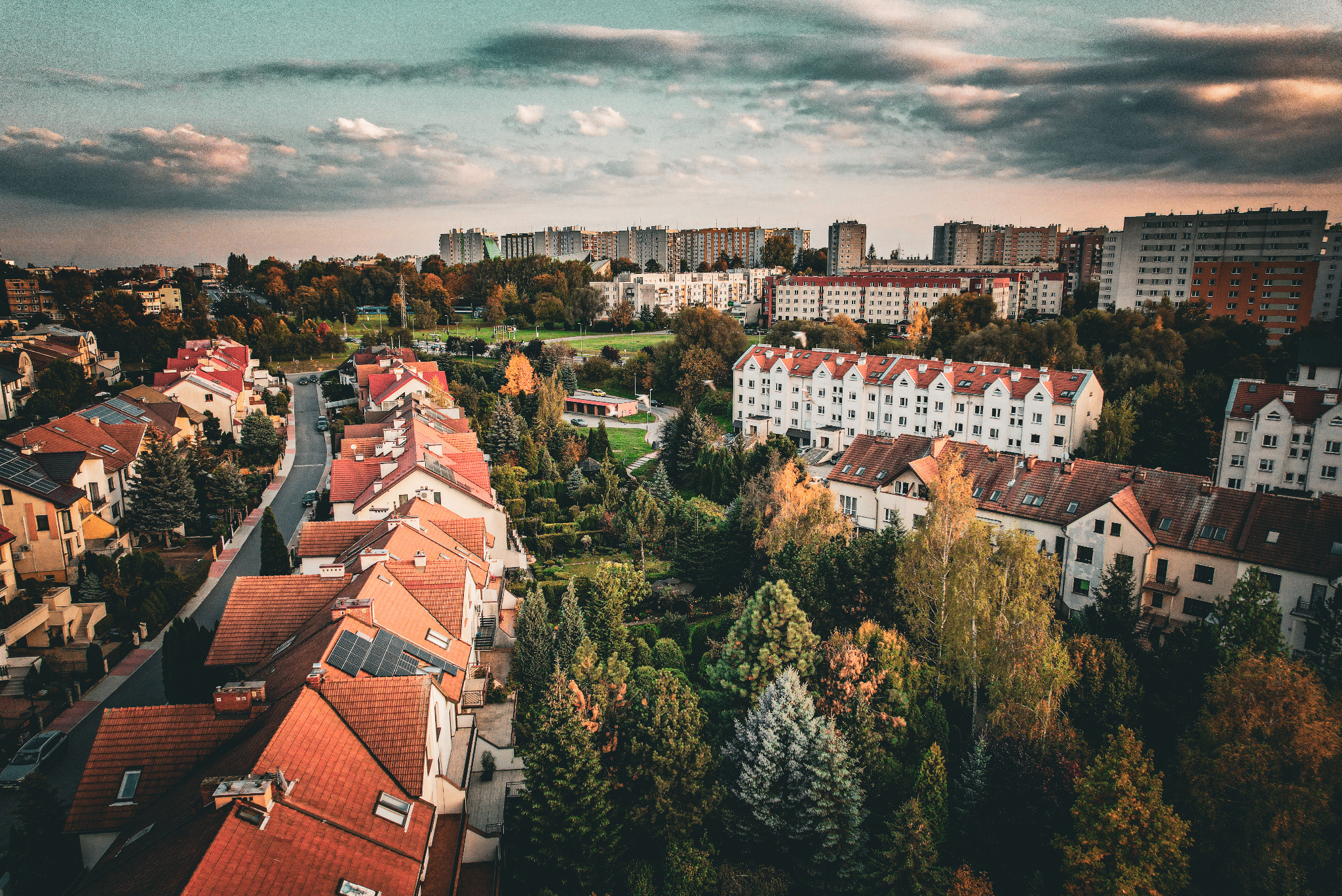 Panoramiczny widok z drona na osiedle domów i bloków, z dominującą czerwoną dachówką i zielenią drzew, pod pochmurnym niebem o zachodzie słońca. Widoczne panele słoneczne na dachach.