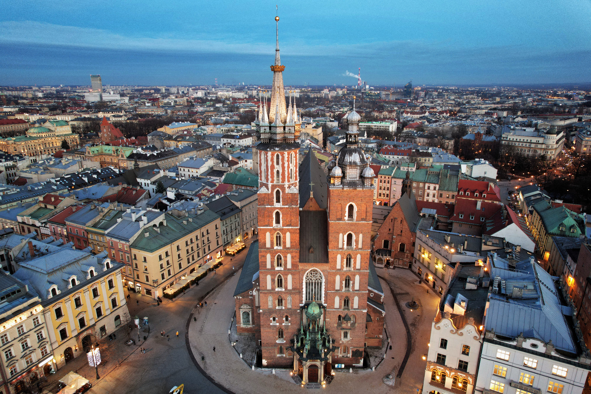 Panorama Krakowa z lotu ptaka o zmierzchu, z Bazyliką Mariacką na pierwszym planie. Widoczne liczne budynki, ulice i oddalony horyzont miasta pod błękitnym niebem.