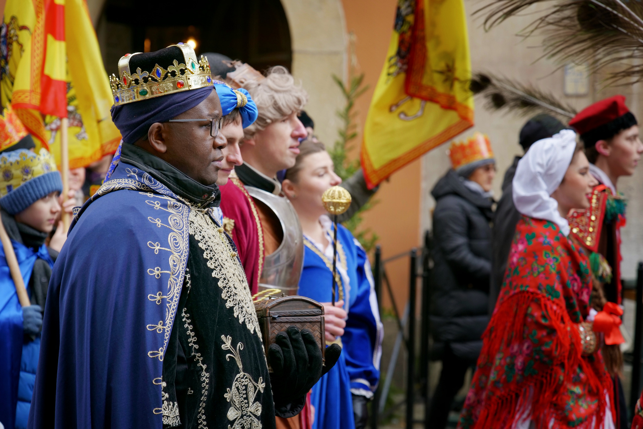 Orszak Trzech Króli w barwnych strojach historycznych, z flagami i rekwizytami, uchwycony z boku podczas przemarszu. Uwagę przykuwa król w koronie i bogatym, zdobionym płaszczu.