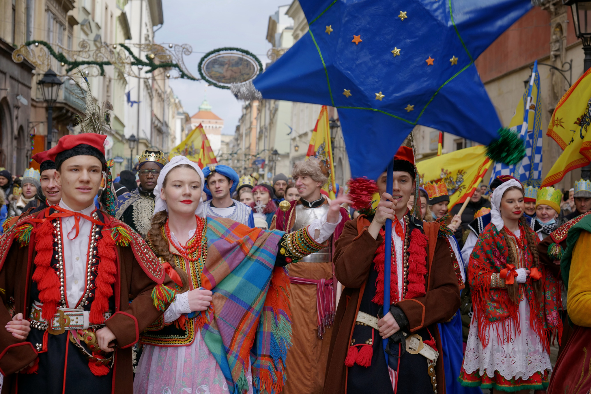 Barwny korowód w tradycyjnych strojach ludowych podczas ulicznego wydarzenia. Uczestnicy niosą flagi i gwiazdę, tworząc radosną atmosferę.