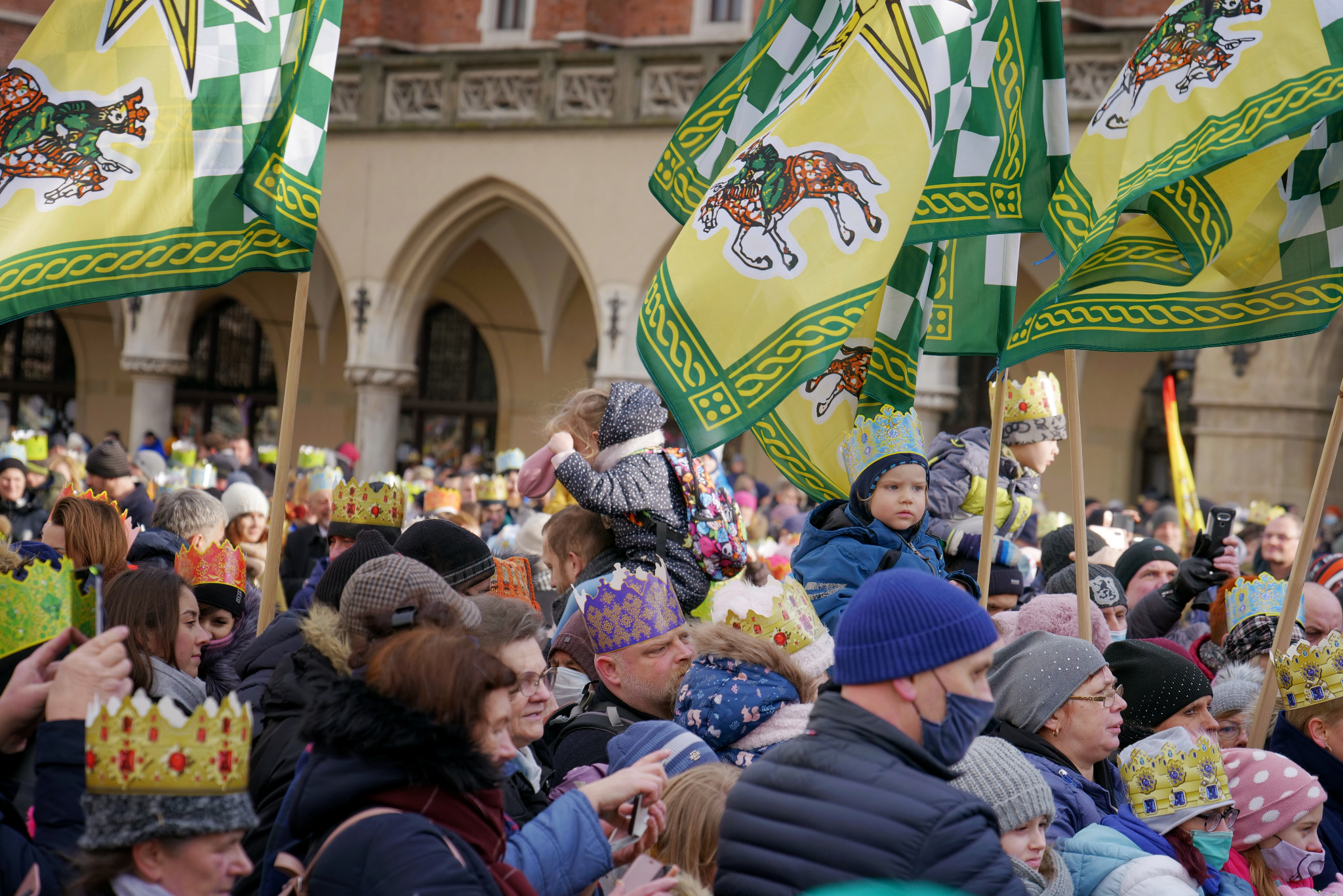 Tłum ludzi w koronach i z flagami podczas Orszaku Trzech Króli na tle zabytkowej architektury. Dziecko na ramionach. Uczestnicy robią zdjęcia telefonami.