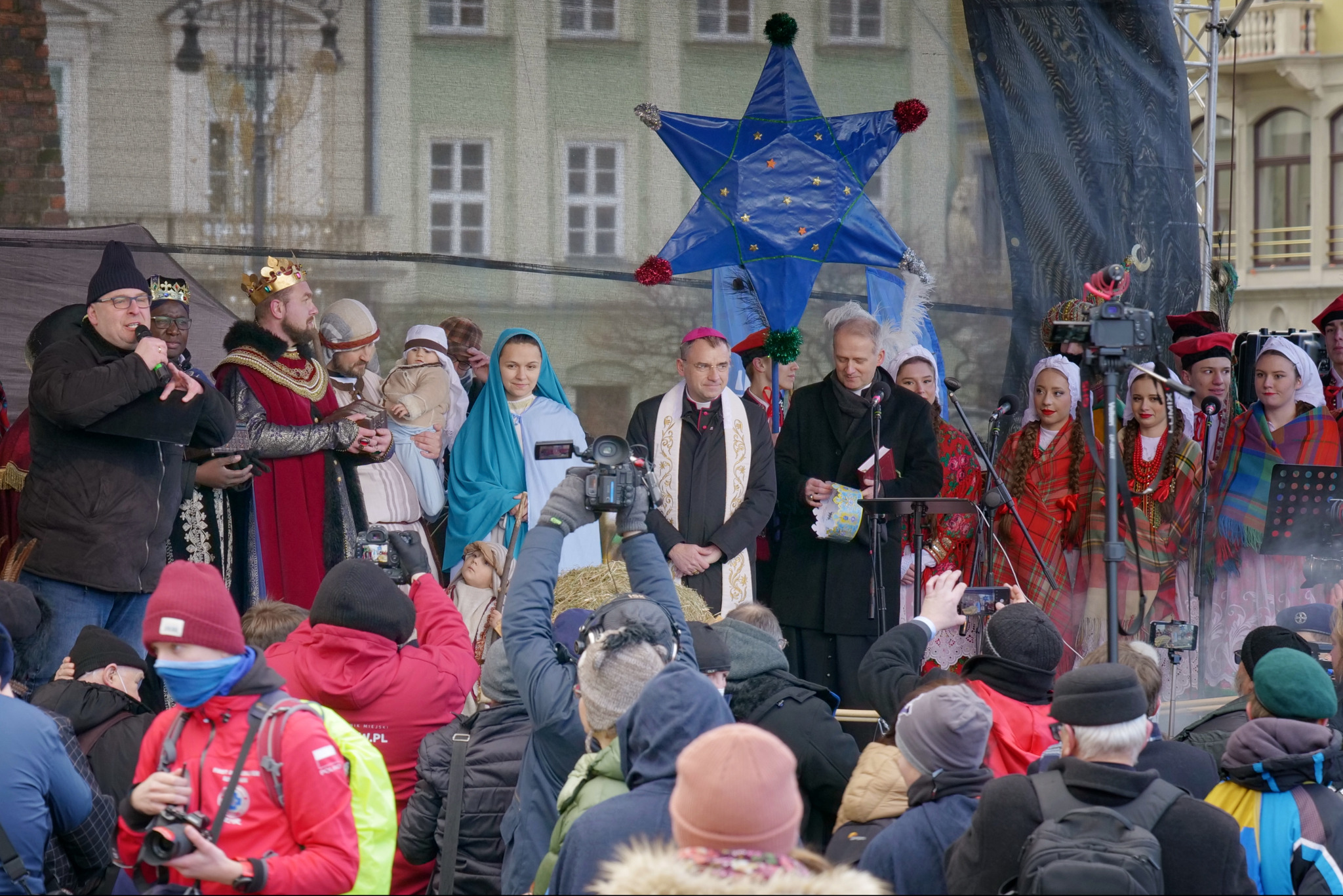 Uroczystość plenerowa z udziałem aktorów w kostiumach, duchownego i publiczności. W tle budynek i dekoracja w kształcie gwiazdy. Widoczni fotografowie i kamerzyści.