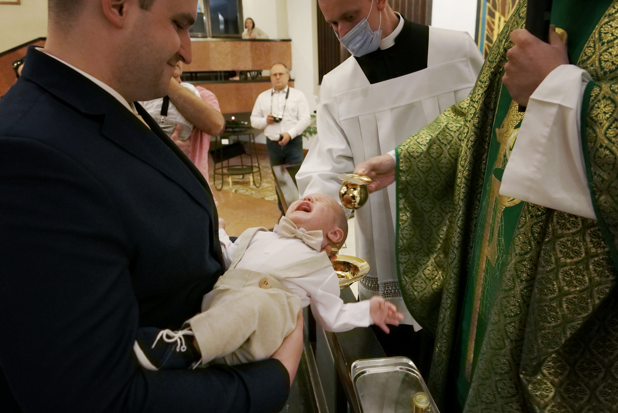 Uśmiechnięty mężczyzna trzyma płaczące dziecko w eleganckim ubranku podczas ceremonii chrztu. W tle ksiądz w masce i osoba z aparatem.