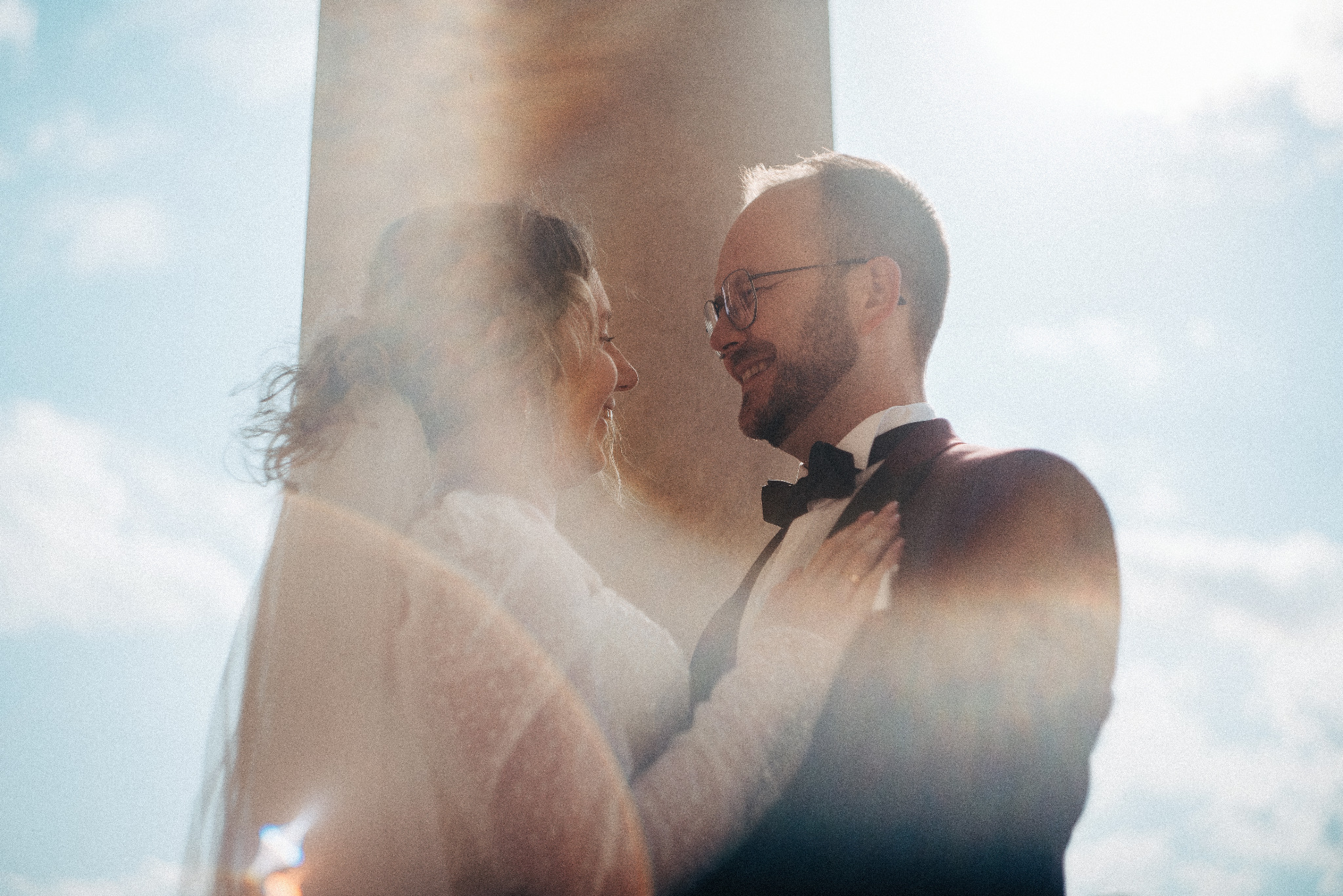 Para młoda w objęciach, on w bordowym smokingu i muszce, ona w białej sukni z welonem, oboje uśmiechnięci, w tle kolumna i błękitne niebo. Romantyczna sesja ślubna w plenerze.