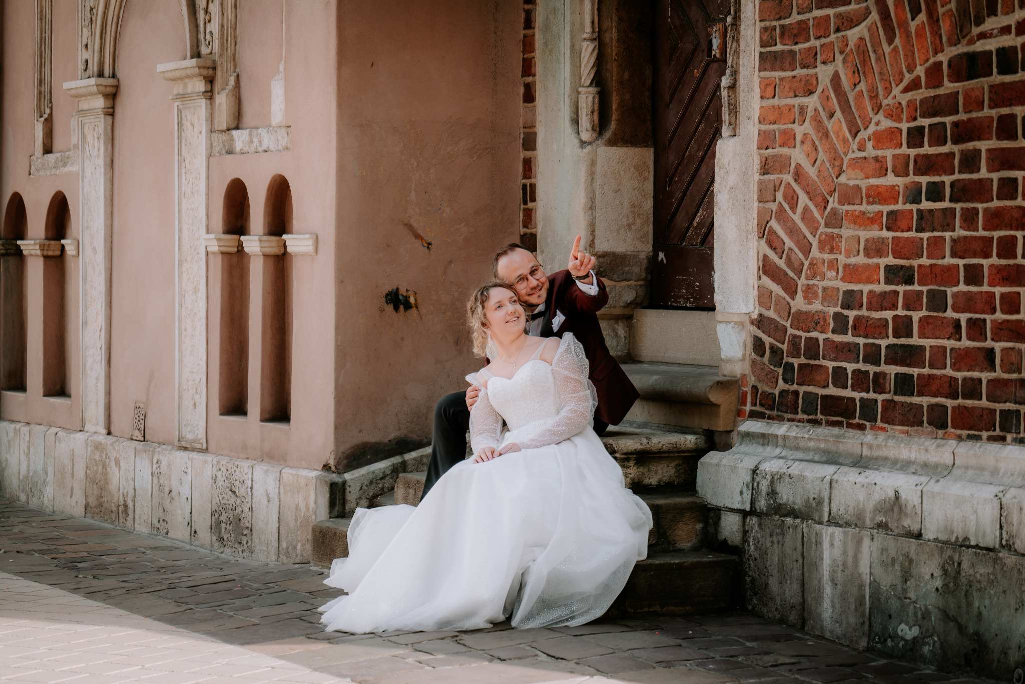 Uśmiechnięta para młoda siedzi na schodach zabytkowej budowli. Pan młody w bordowym garniturze wskazuje palcem w górę. Romantyczna sesja ślubna w plenerze.