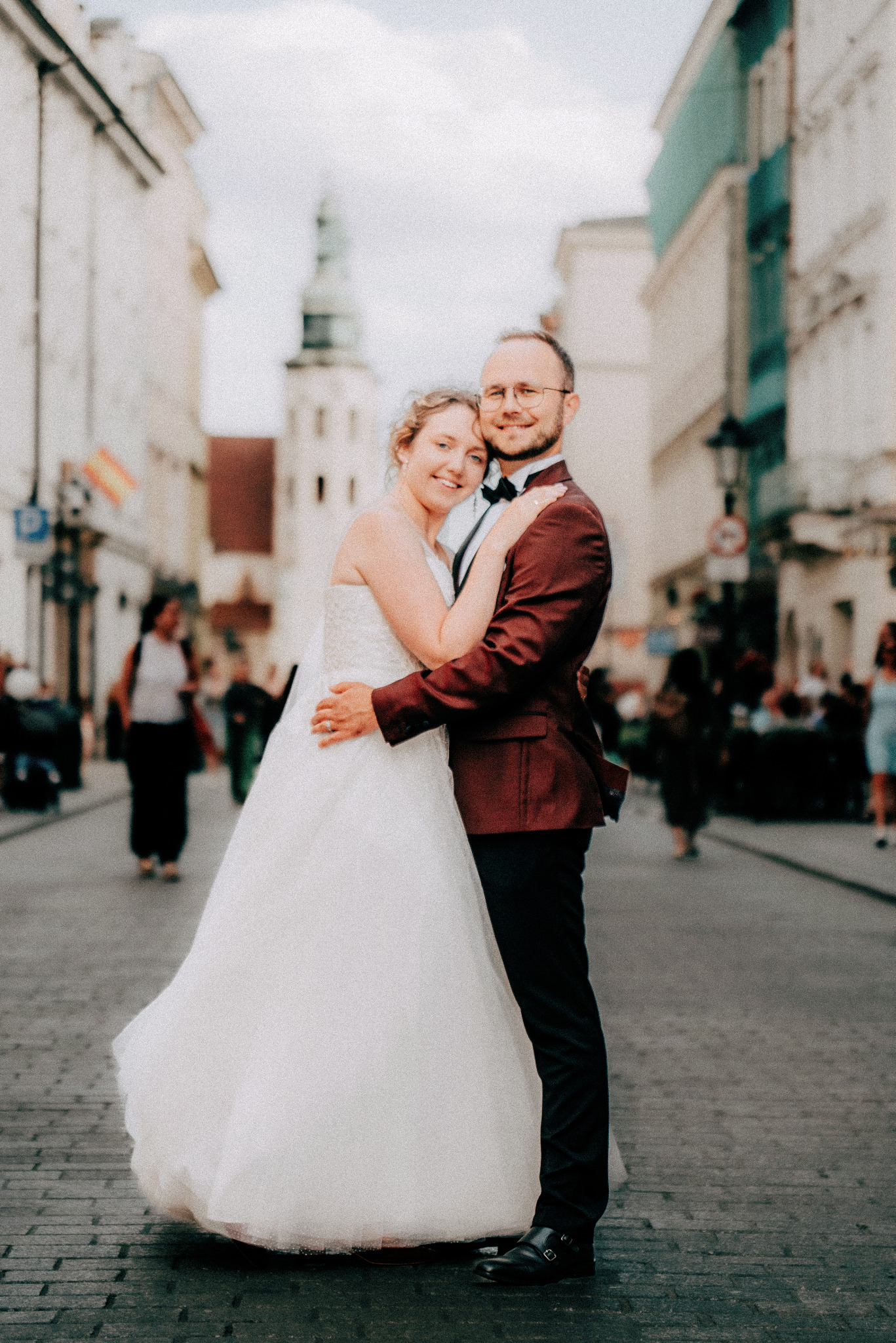 Uśmiechnięta para młoda w objęciach na tle brukowanej ulicy i zabytkowej architektury. Pan w bordowej marynarce, pani w białej sukni ślubnej. Romantyczna sesja plenerowa.