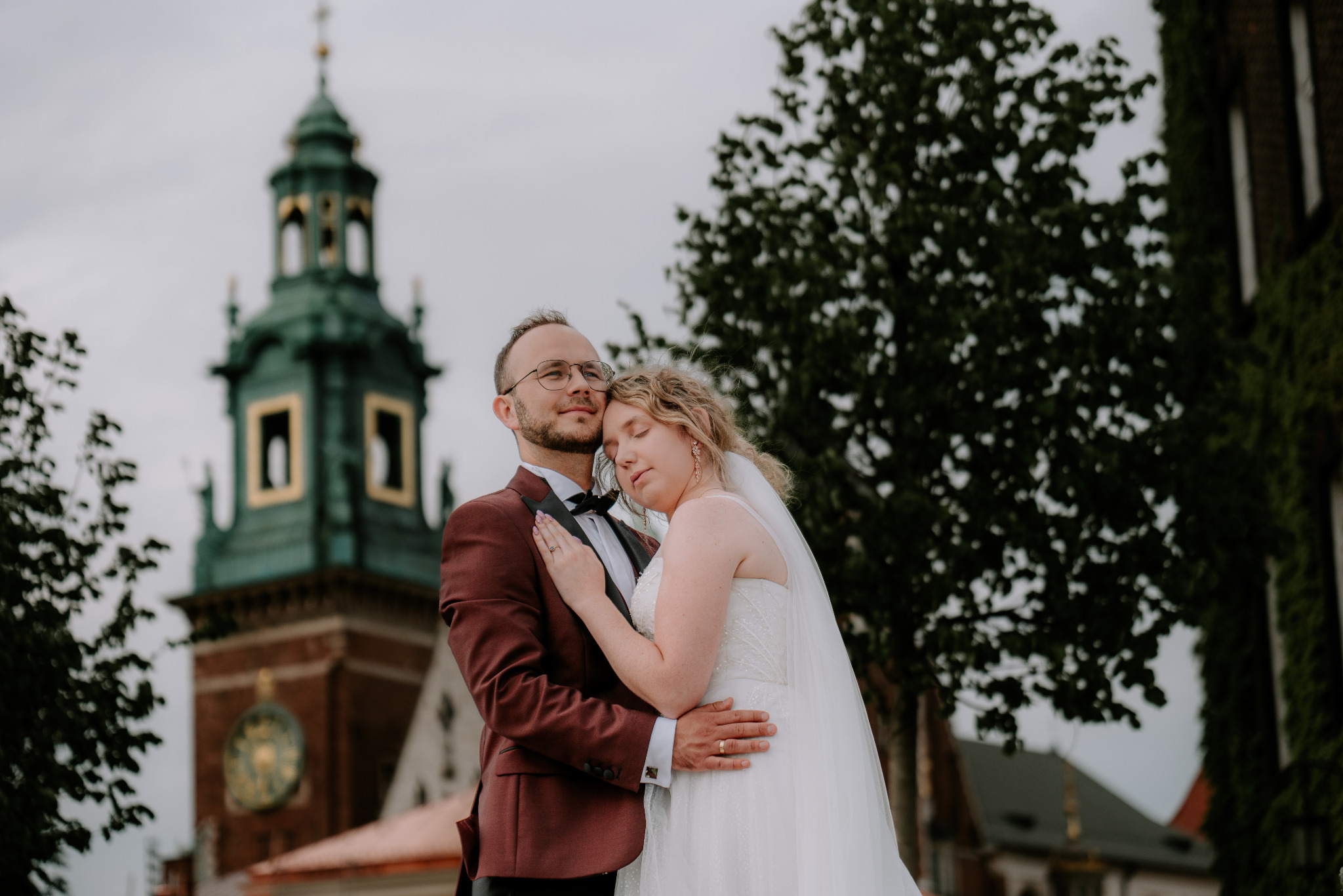 Para młoda w objęciach na tle Wawelu. Pan młody w bordowym smokingu, panna młoda w białej sukni. Romantyczna sesja ślubna w Krakowie.