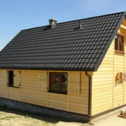 Gotowy domek letniskowy z jasnego drewna z czarnym dachem w Dukli, z widocznymi drewnianymi okiennicami i fragmentem tarasu.