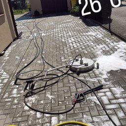 Electrical Cleaner - Czyszczenie nawierzchni z kostki brukowej za pomocą profesjonalnej myjki powierzchniowej, z widocznymi wężami i pianą. W tle budynek z panelami słonecznymi.