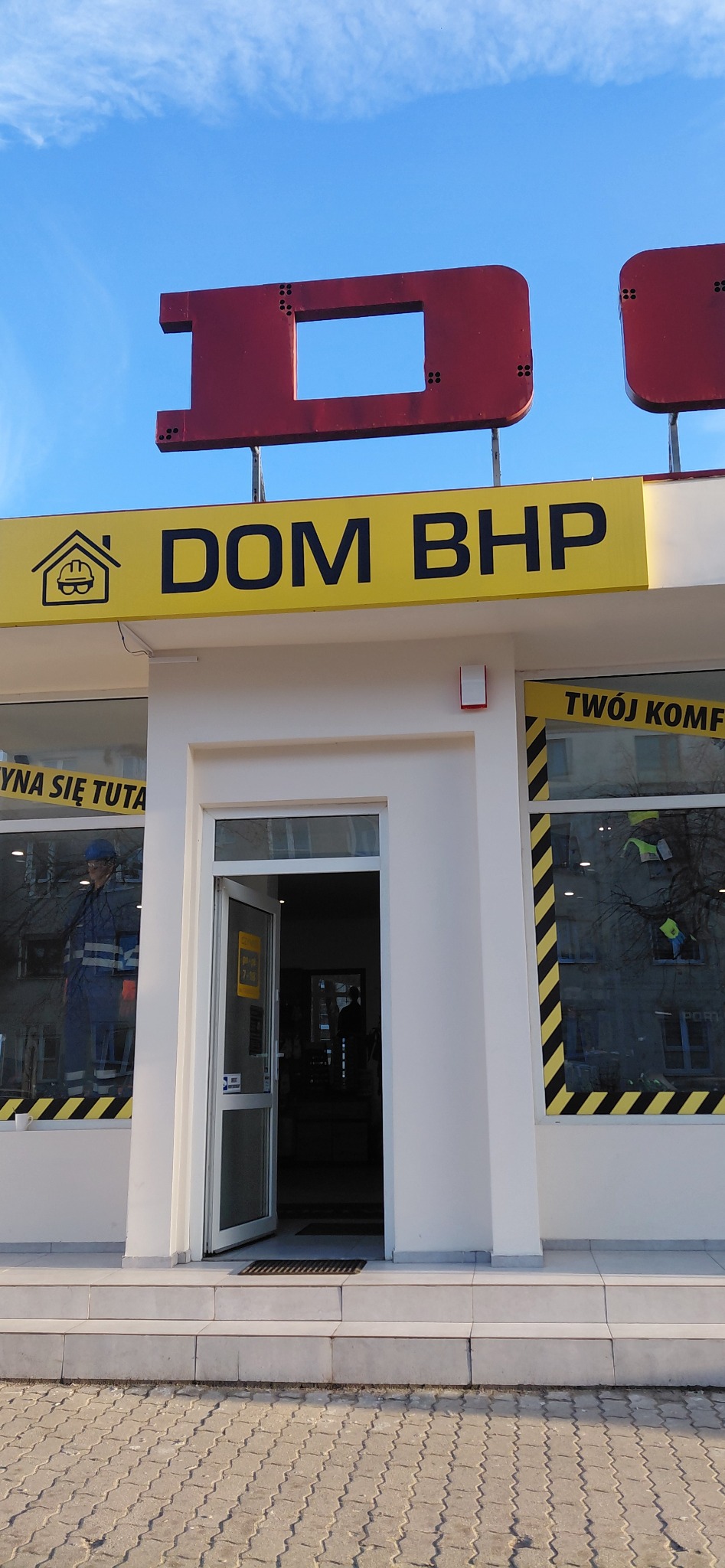 Fasada sklepu Dom BHP z manekinem w odzieży roboczej w oknie. Widoczny szyld z logo i otwarte drzwi wejściowe. Błękitne niebo w tle.