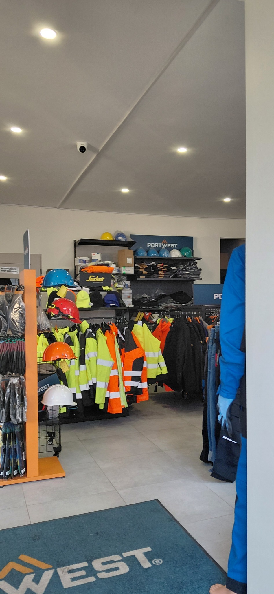 Wnętrze sklepu z odzieżą roboczą: kaski, kamizelki odblaskowe i ubrania ochronne na wieszakach. Widoczny fragment maty podłogowej z logo firmy.