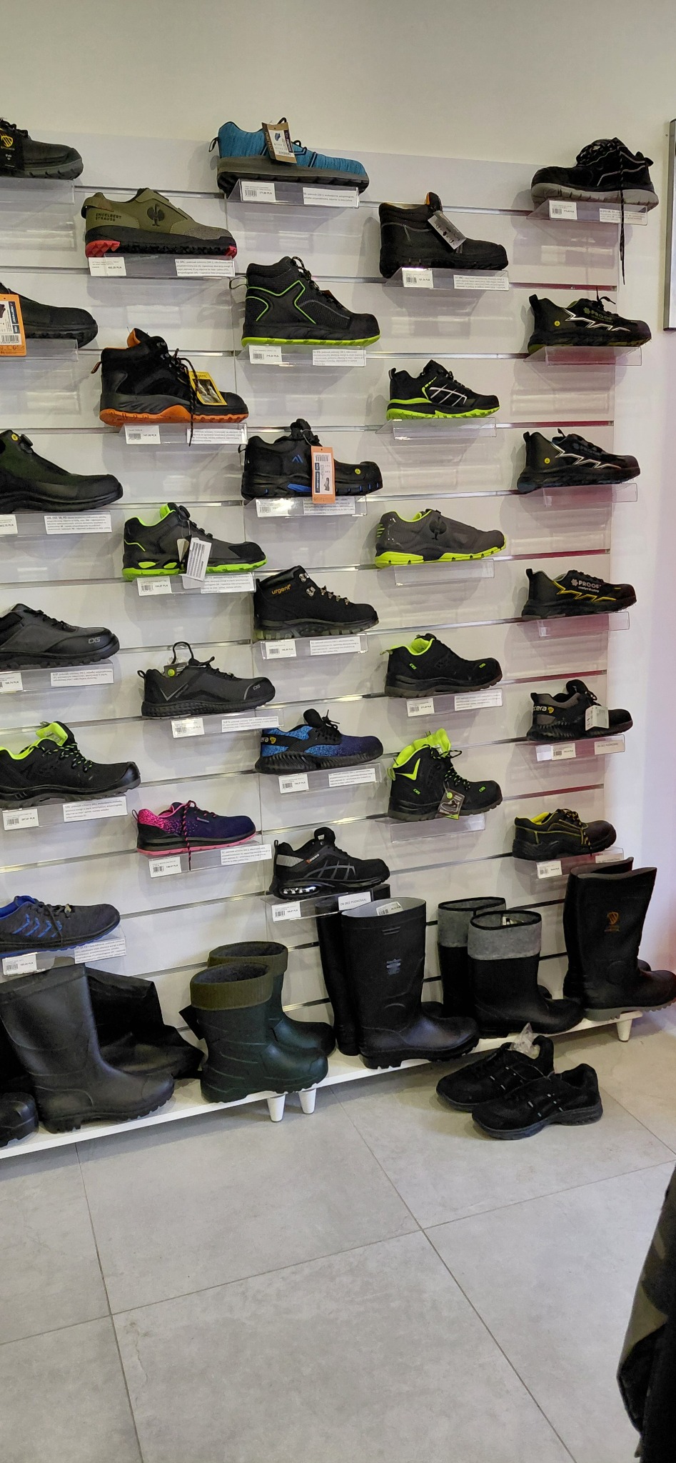 Ekspozycja obuwia roboczego na półkach w sklepie. Różne modele butów, w tym wysokie gumowce i niskie sportowe, z widocznymi cenami i metkami.