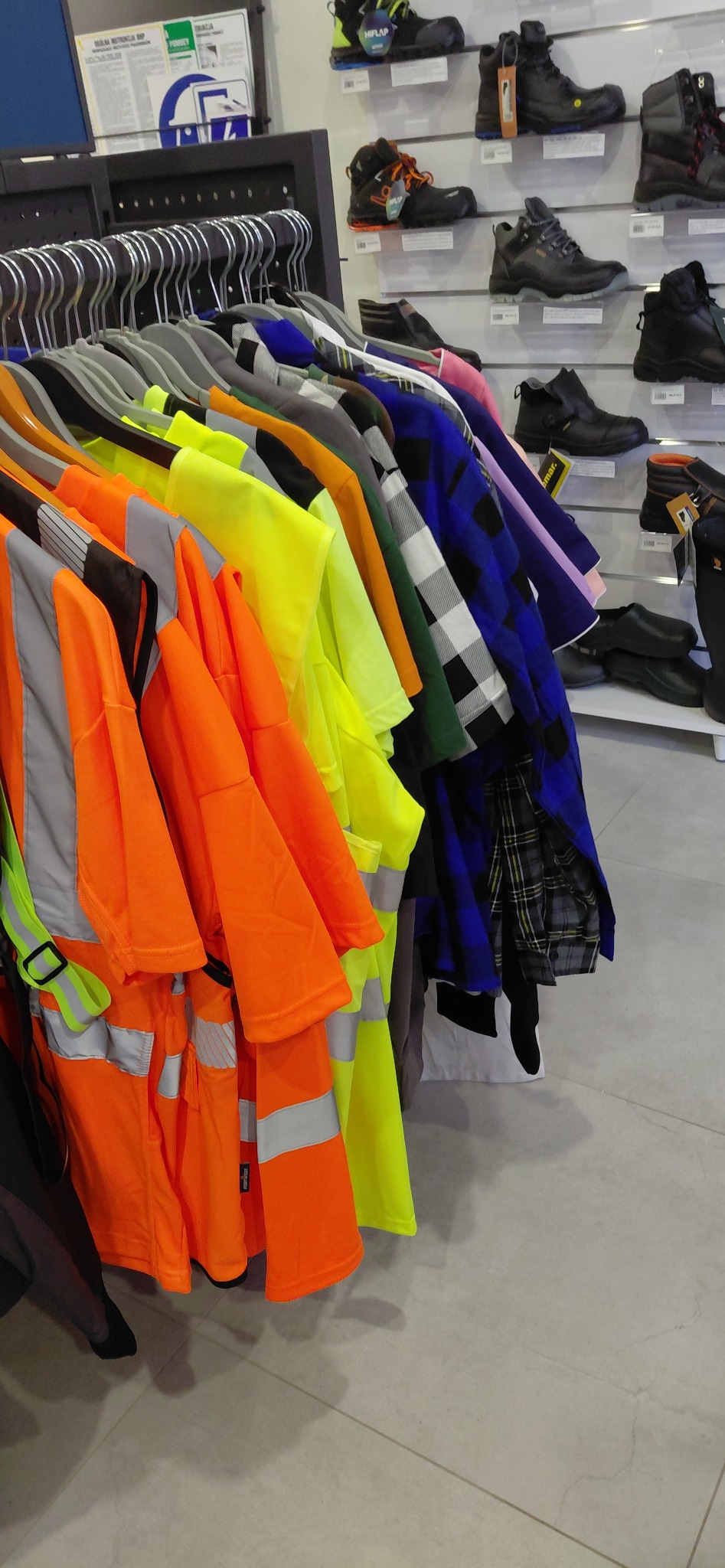 Różnorodna odzież robocza na wieszakach: pomarańczowe kamizelki odblaskowe, neonowe koszulki i flanelowe koszule w kratę. W tle buty robocze na półkach.