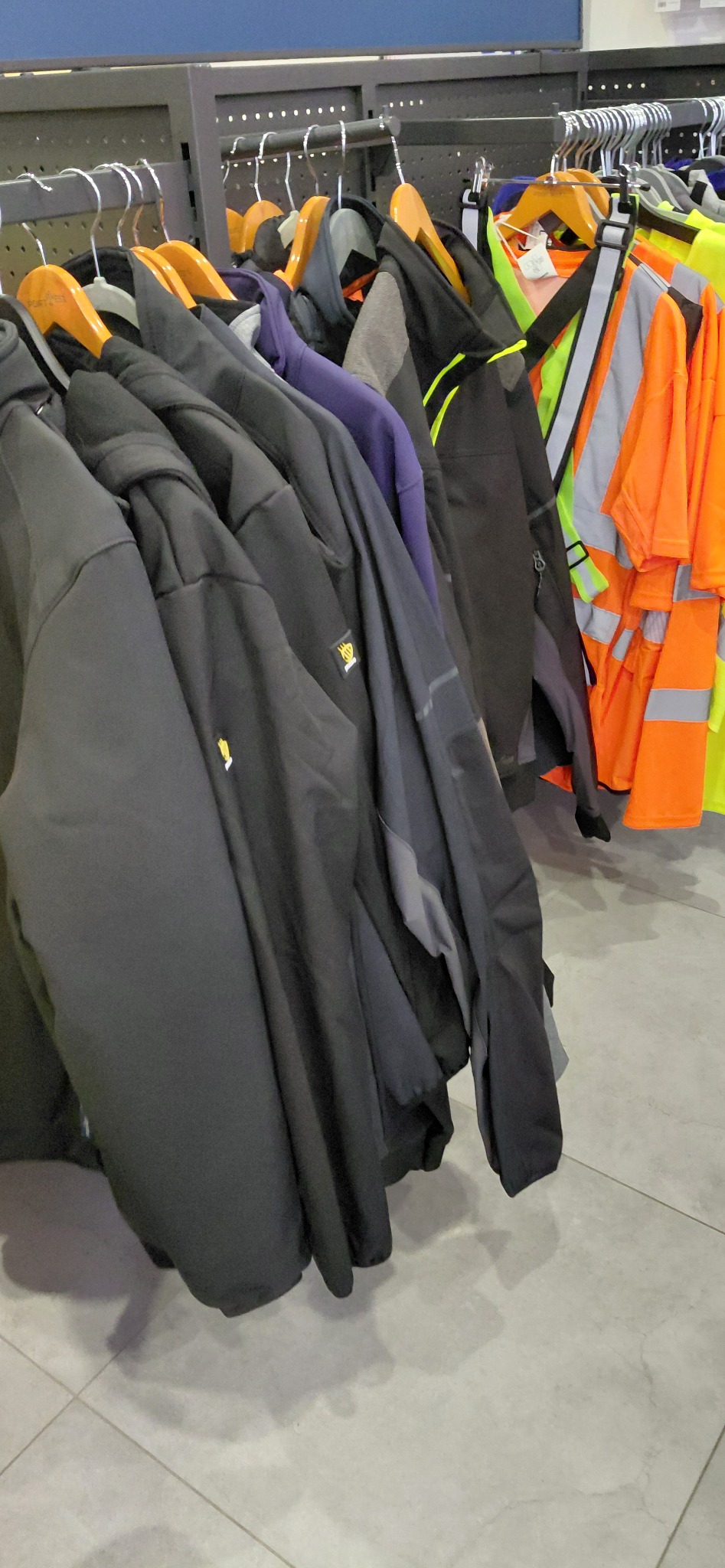 Rząd wieszaków z kurtkami roboczymi, w tym czarne softshelle z logo i pomarańczowe kamizelki odblaskowe, na tle szarej podłogi i ciemnej ściany.