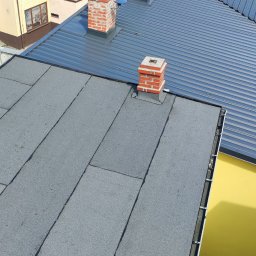 MBud Michał Naciskała - Widok z góry na dach z papy i blachy trapezowej, z ceglanymi kominami. Dach sąsiaduje z żółtą ścianą budynku. Na pierwszym planie komin z drabinką.