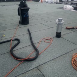 MBud Michał Naciskała - Szary dach pokryty papą termozgrzewalną z widocznymi narzędziami: wąż, szczotka, butla gazowa i pojemnik na odpady. W tle worki z materiałami budowlanymi.