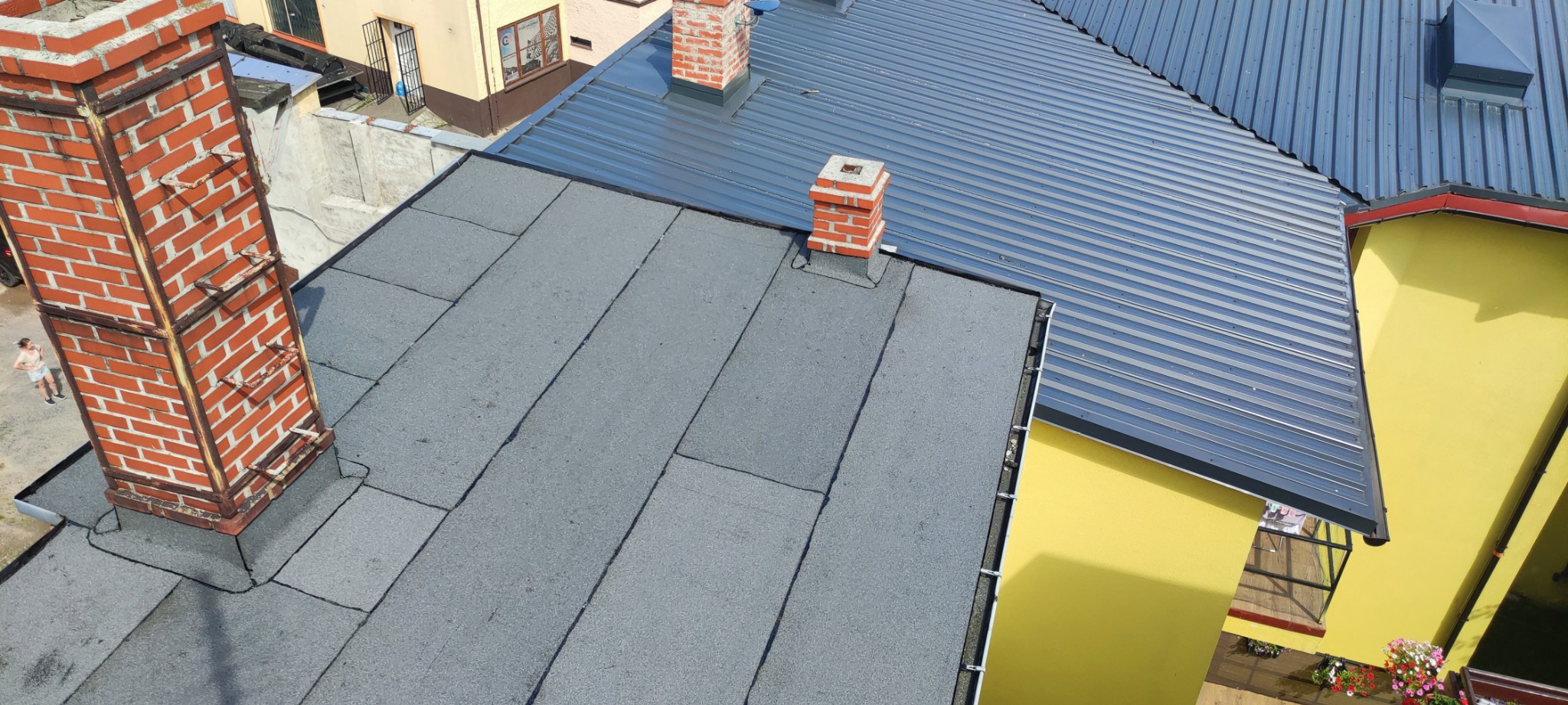 Widok z góry na dach z papy i blachy trapezowej, z ceglanymi kominami. Dach sąsiaduje z żółtą ścianą budynku. Na pierwszym planie komin z drabinką.