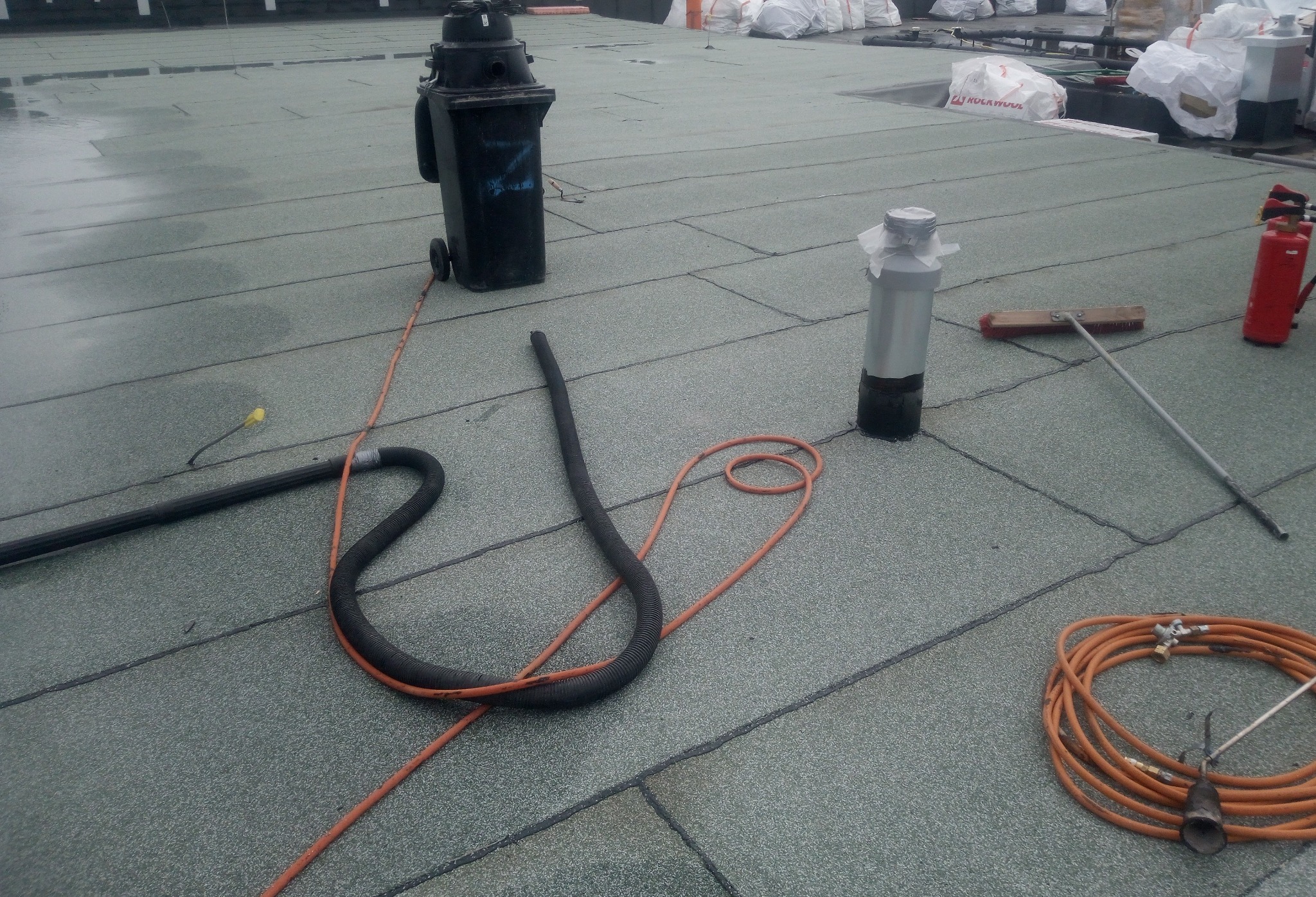 Szary dach pokryty papą termozgrzewalną z widocznymi narzędziami: wąż, szczotka, butla gazowa i pojemnik na odpady. W tle worki z materiałami budowlanymi.