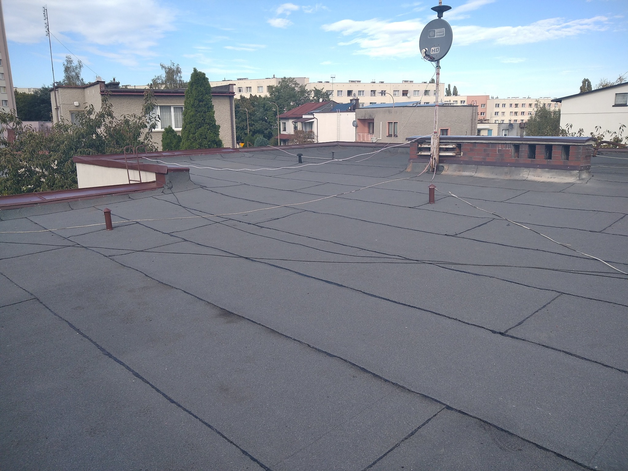 Płaski dach pokryty papą termozgrzewalną, widoczne kominy i instalacje antenowe na tle zabudowy miejskiej w słoneczny dzień. Charakterystyczne łączenia papy.