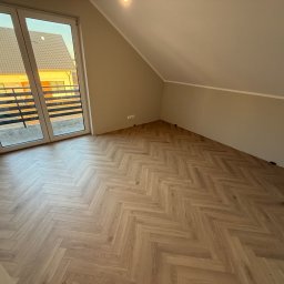 Mateusz Pikul - Jasne panele ułożone w jodełkę w pokoju na poddaszu z oknem balkonowym. Ściany w odcieniach beżu, widoczne listwy przypodłogowe i gniazdka elektryczne. Nowoczesne wykończenie.