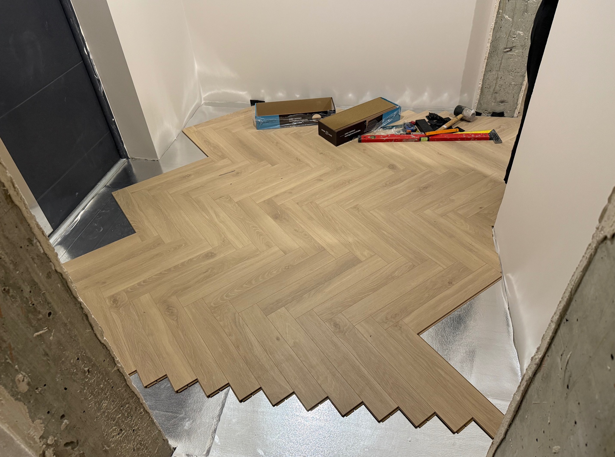 Fragment układania jasnych paneli podłogowych w jodełkę. Widoczne narzędzia, podkład i niedokończony fragment podłogi w pomieszczeniu w trakcie prac wykończeniowych.