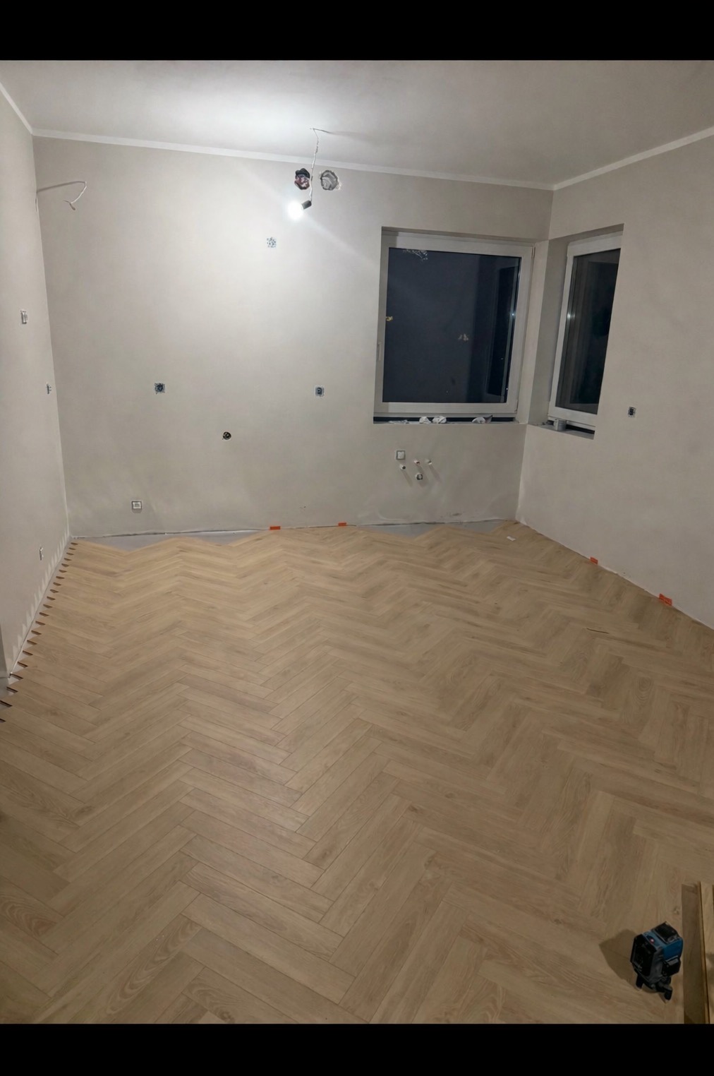 Jasne panele ułożone w jodełkę w pokoju z dwoma oknami. Widoczne listwy przypodłogowe i poziomica laserowa. Ściany przygotowane do malowania.