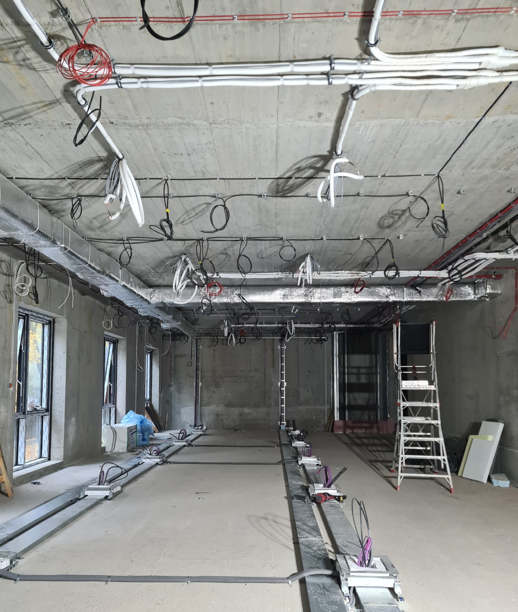 Wnętrze w stanie surowym z widoczną instalacją elektryczną na suficie i podłodze. Kable i przewody ułożone w korytach, drabina aluminiowa w tle. Widoczne okna i surowy beton.