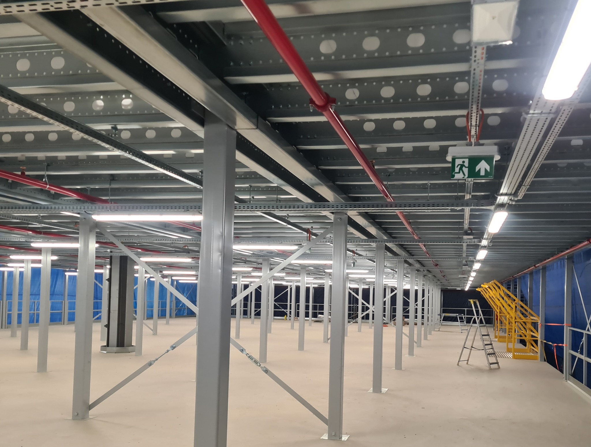 Wnętrze hali magazynowej z metalową konstrukcją wsporczą, instalacją elektryczną i przeciwpożarową. Widoczne oświetlenie LED i drabiny. Hala w trakcie budowy lub wykańczania.