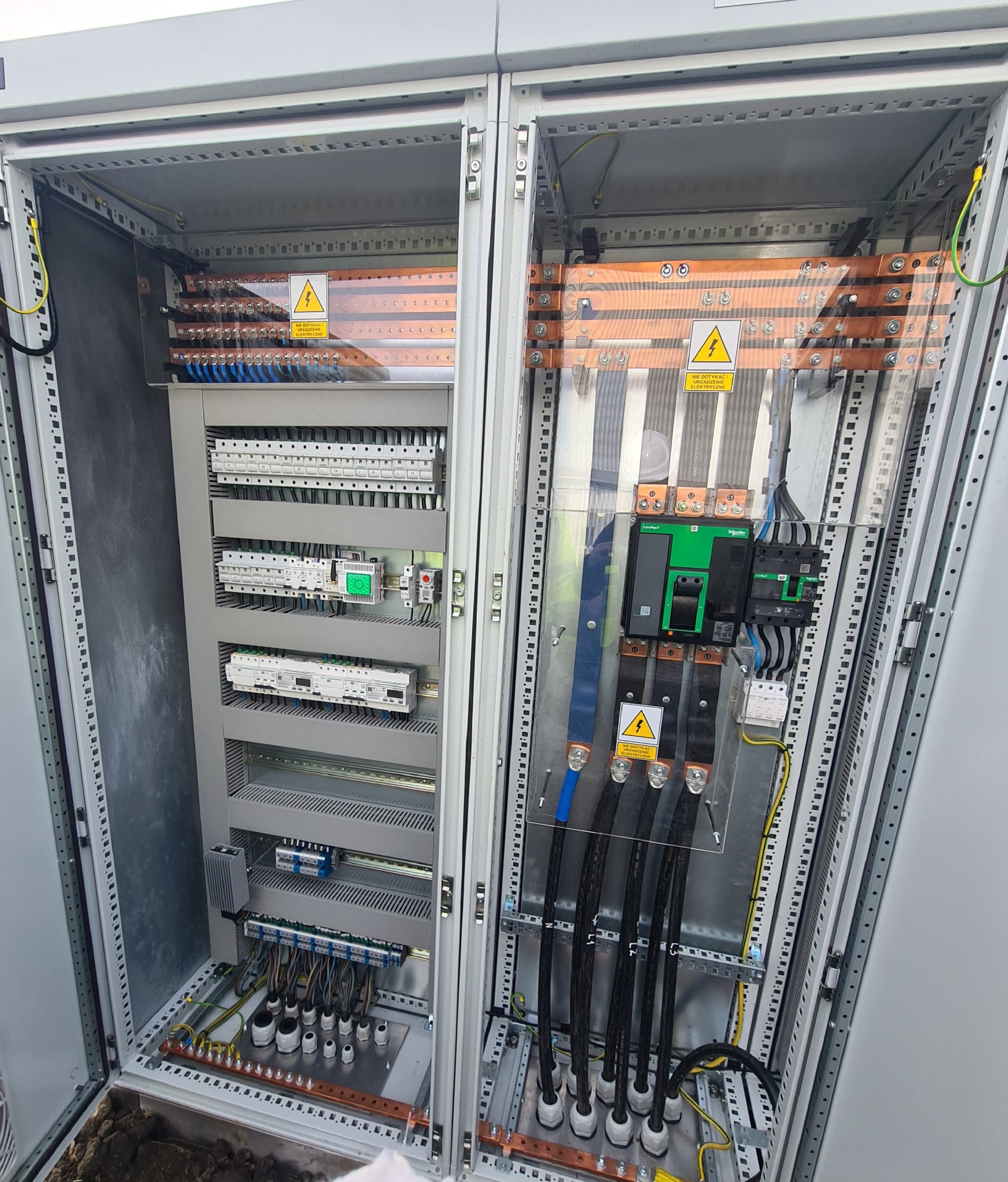 Otwarta szafa elektryczna z widocznymi bezpiecznikami, przewodami i elementami instalacji. Tabliczki ostrzegawcze o napięciu. Widok z bliska na wnętrze rozdzielnicy.