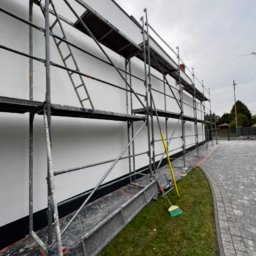 Dene spółka z ograniczoną odpowiedzialnością - Biała elewacja domu w trakcie prac wykończeniowych, widoczne rusztowania, wiadro z farbą i szczotka. Ujęcie z dołu, trawnik i kostka brukowa w tle.