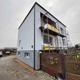 Dene spółka z ograniczoną odpowiedzialnością - Nowoczesny dom w trakcie ocieplania elewacji, widoczne rusztowania i materiały izolacyjne. Dolna część budynku pomalowana na szaro, górna w jasnym odcieniu bieli.