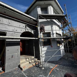 Dene spółka z ograniczoną odpowiedzialnością - Dom w trakcie ocieplania: część elewacji pokryta szarym styropianem, druga w surowym tynku. Metalowe rusztowanie przy ścianie. Widoczne zabezpieczenia na oknach.