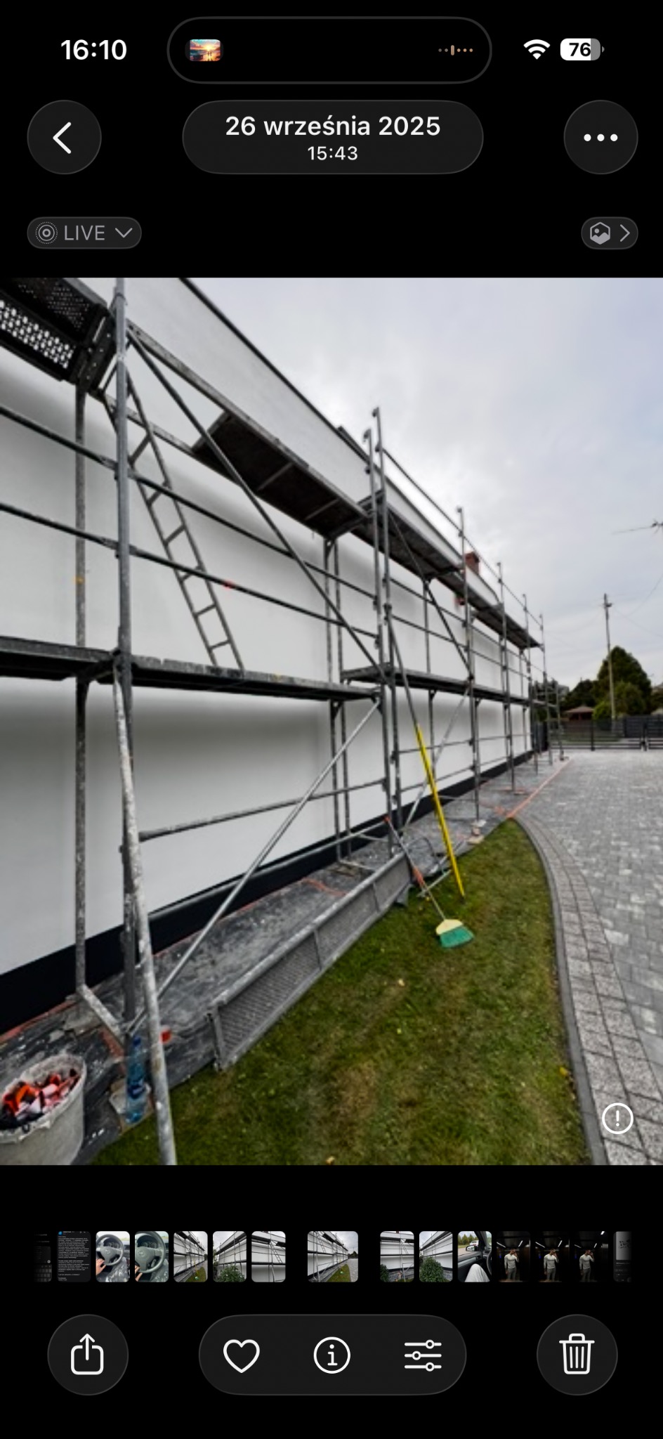 Biała elewacja domu w trakcie prac wykończeniowych, widoczne rusztowania, wiadro z farbą i szczotka. Ujęcie z dołu, trawnik i kostka brukowa w tle.