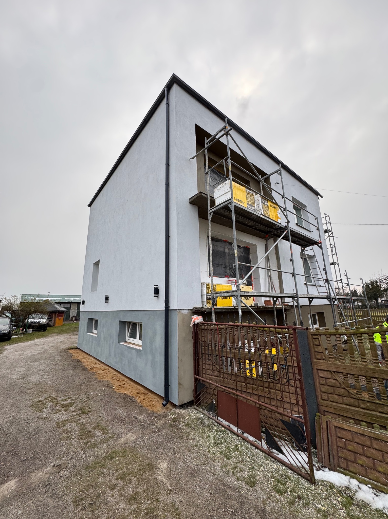 Nowoczesny dom w trakcie ocieplania elewacji, widoczne rusztowania i materiały izolacyjne. Dolna część budynku pomalowana na szaro, górna w jasnym odcieniu bieli.