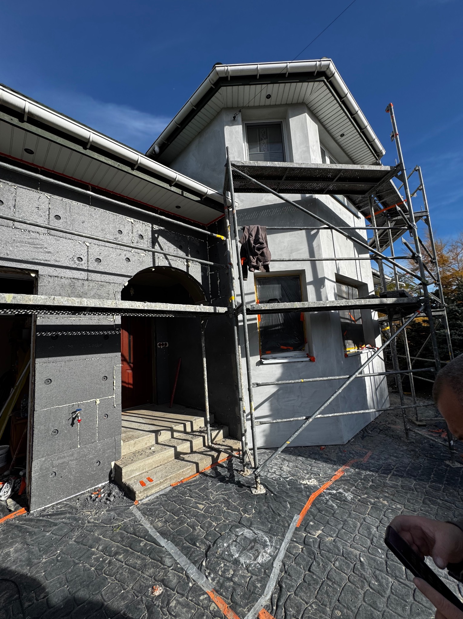 Dom w trakcie ocieplania: część elewacji pokryta szarym styropianem, druga w surowym tynku. Metalowe rusztowanie przy ścianie. Widoczne zabezpieczenia na oknach.