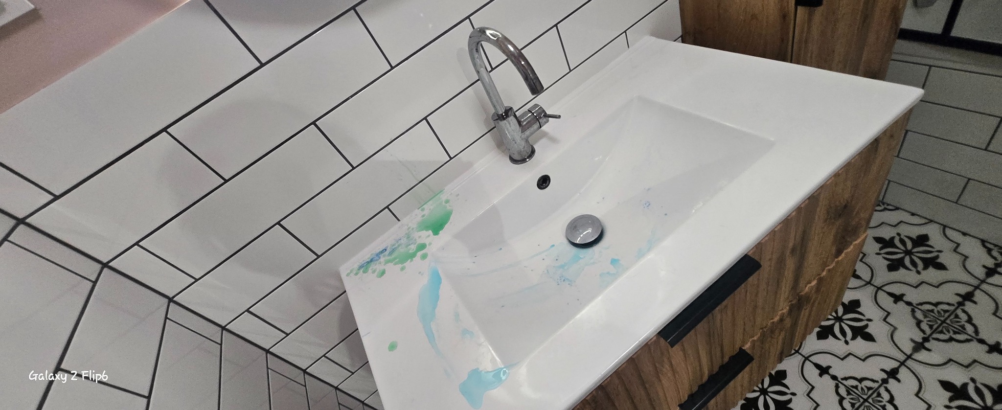 Zabrudzona umywalka z plamami niebieskiego i zielonego płynu, chromowana bateria, białe płytki na ścianie i wzorzysta podłoga. Widok z góry, ukośny kadr.