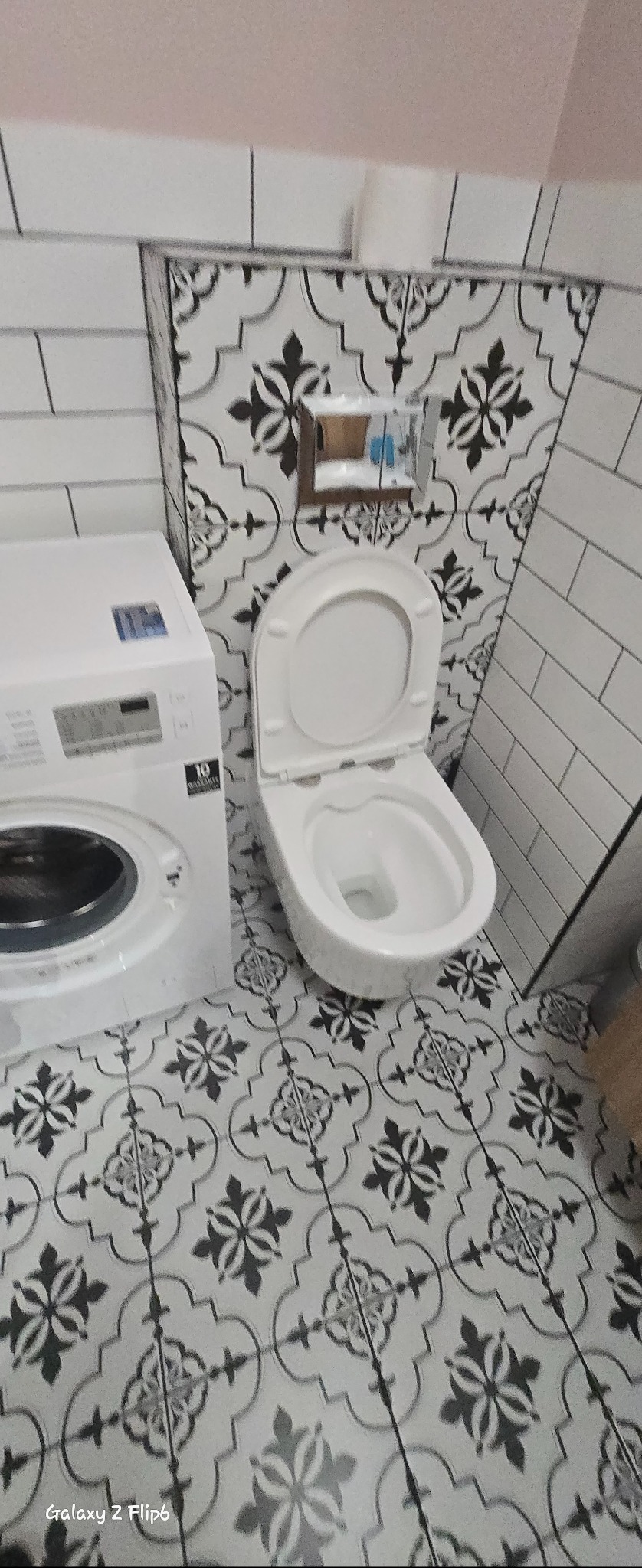 Biała toaleta z otwartą klapą, obok pralka, na podłodze i ścianach wzorzyste płytki w czarno-białe motywy. Widok z góry, ukośny kadr.