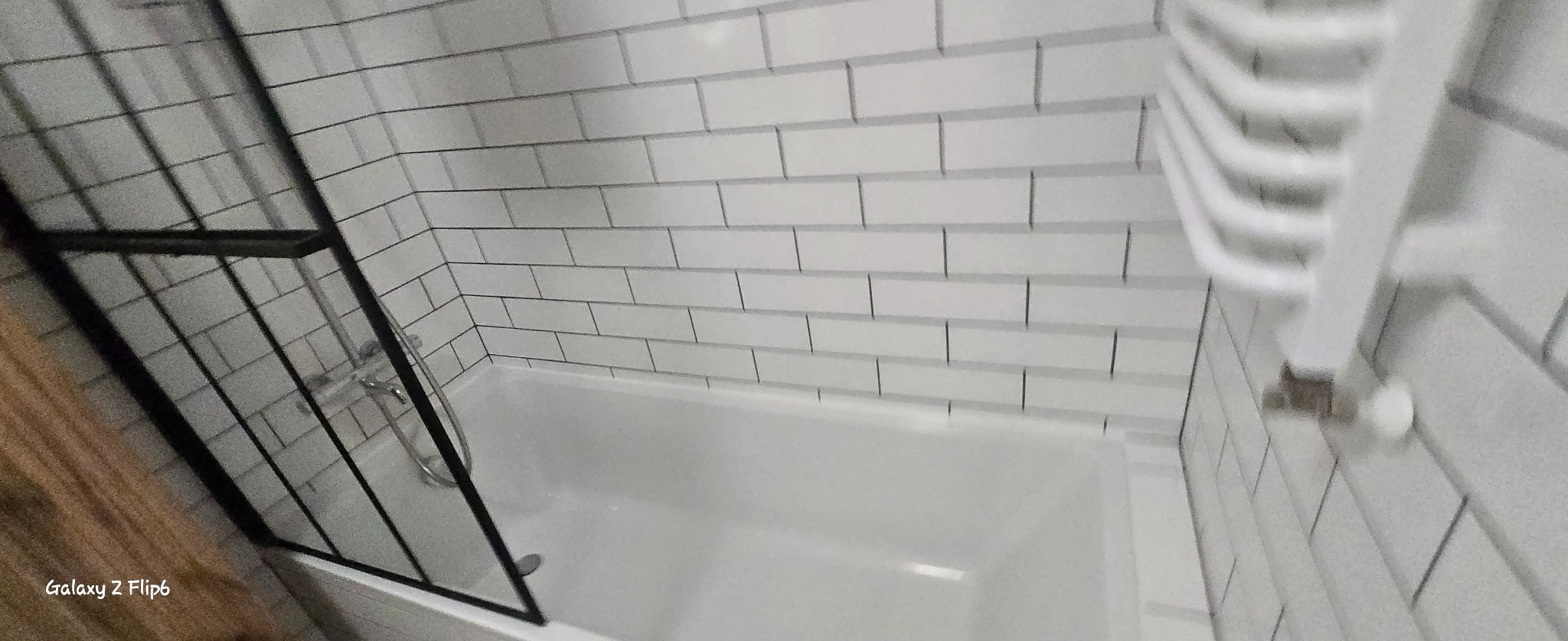 Biała wanna z czarną kabiną prysznicową i grzejnikiem drabinkowym na ścianie z białych płytek w stylu cegiełek. Minimalistyczna łazienka po remoncie.