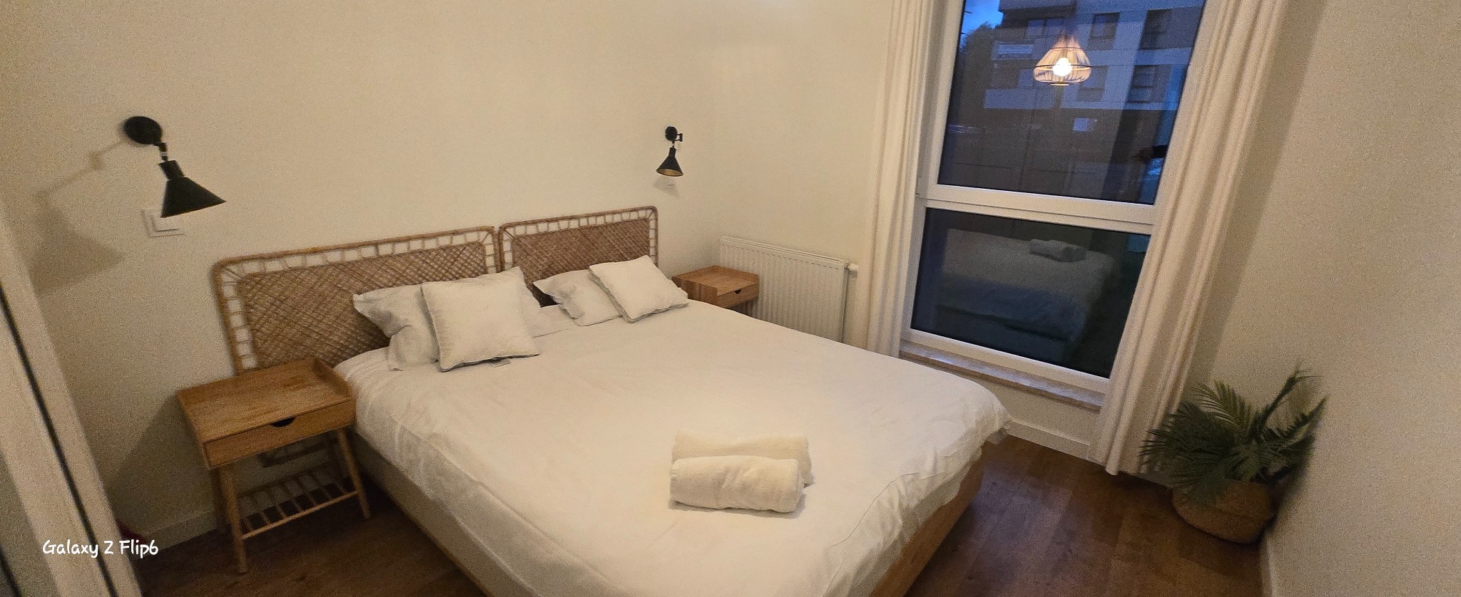 Jasna sypialnia z wiklinowym zagłówkiem, białymi poduszkami i lampkami nocnymi. Widok na budynek zza okna. Minimalistyczny wystrój, czystość i porządek.