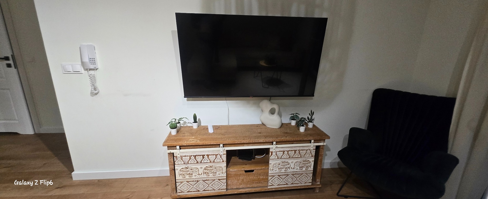 Telewizor na ścianie nad drewnianą komodą z dekoracyjnymi wzorami, obok fotel. Na komodzie rośliny w doniczkach i biała rzeźba. Widok na wnętrze mieszkania.