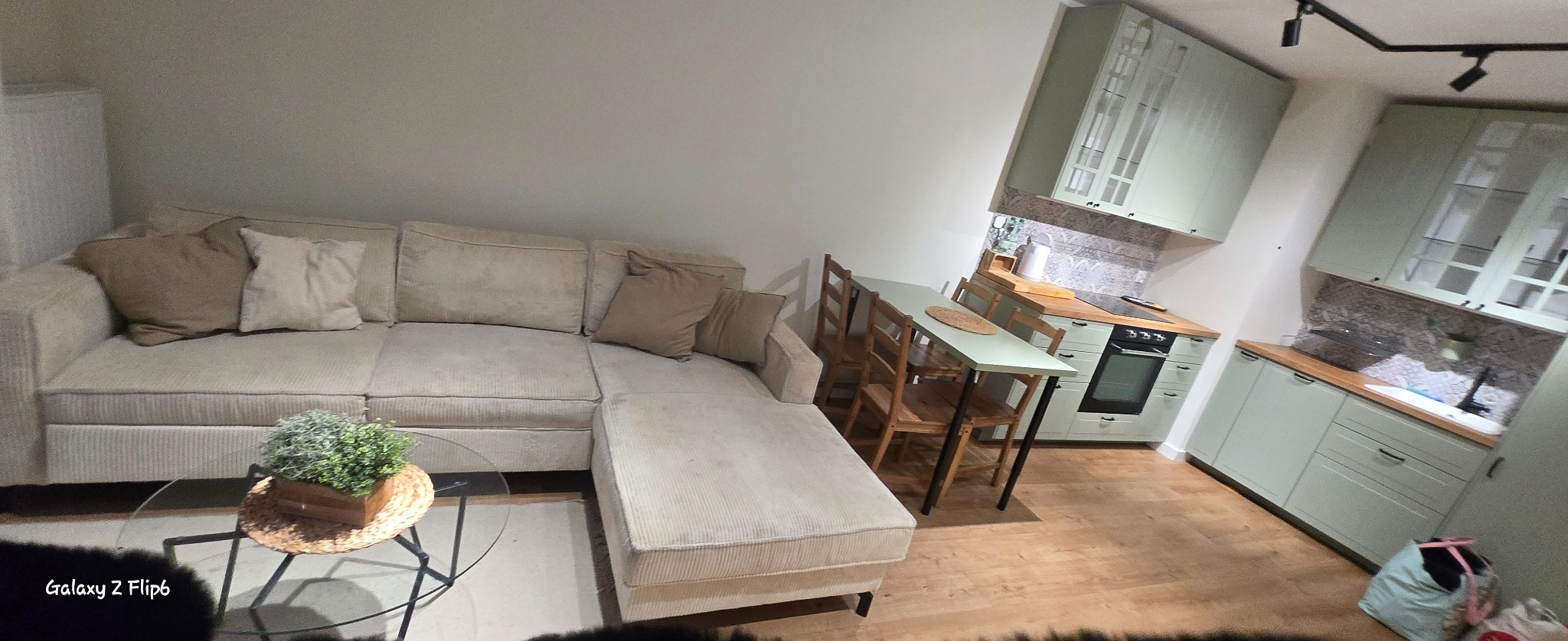 Jasny salon z narożnikiem, stolikiem kawowym i zieloną kuchnią w tle. Widok na wnętrze mieszkania po remoncie, z elementami dekoracyjnymi i meblami.