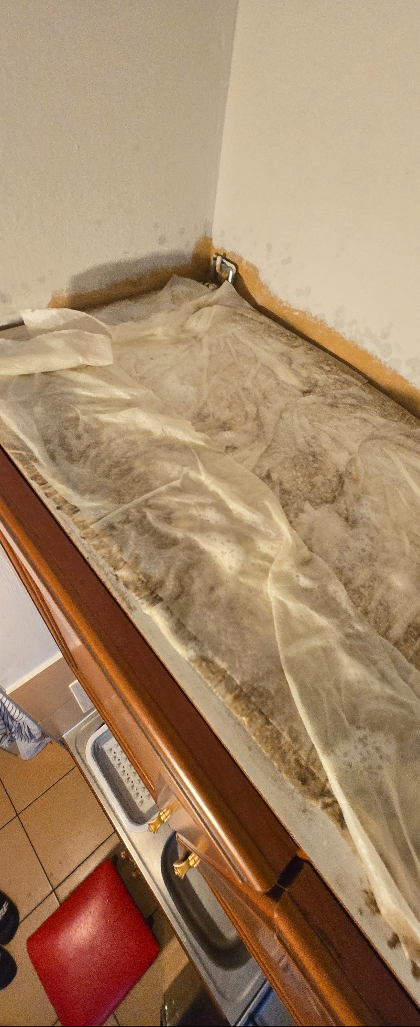 Zabezpieczony blat kuchenny folią ochronną przed remontem. Widoczne ślady wilgoci na ścianie i blacie. Ujęcie z góry, fragment zlewu i czerwonego krzesła.