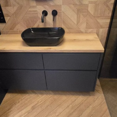 Czarna, owalna umywalka nablatowa na drewnianym blacie szafki w łazience z geometryczną mozaiką na ścianie i podłodze ułożoną w jodełkę.