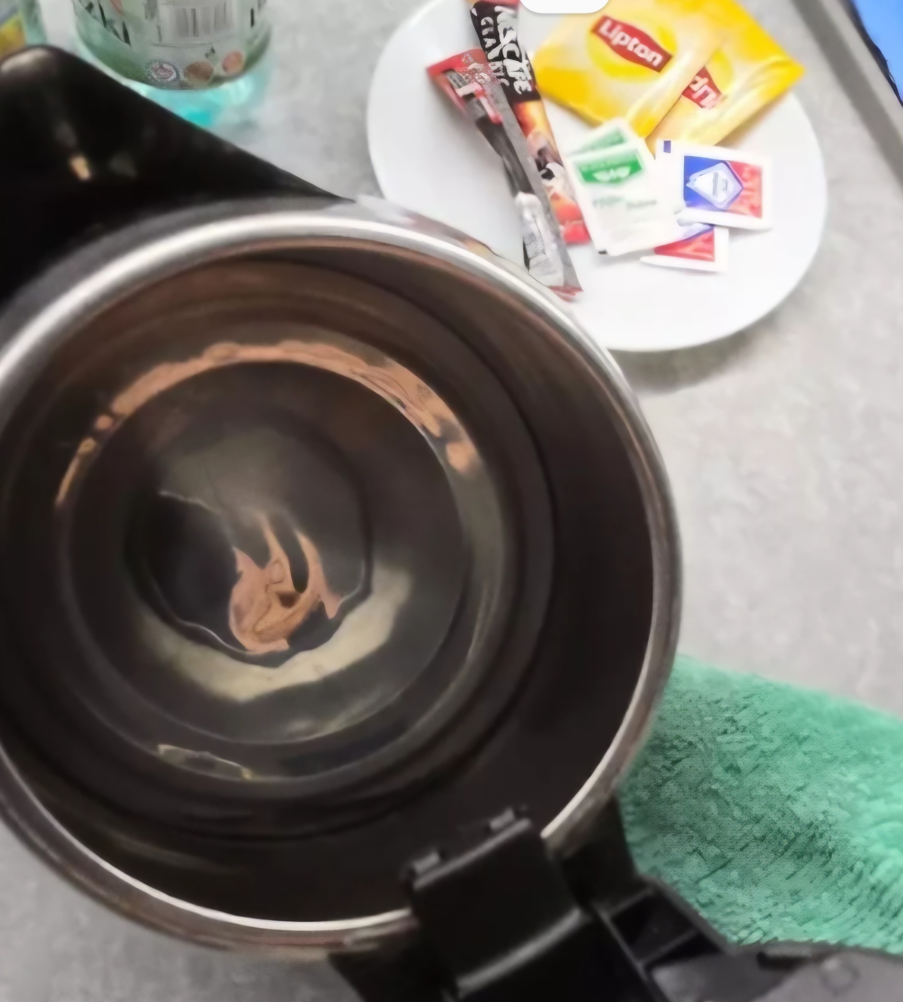 Wnętrze czajnika elektrycznego z osadem, obok saszetki herbaty i kawa na białym talerzyku. Widoczna butelka wody. Perspektywa z góry, bliski kadr.