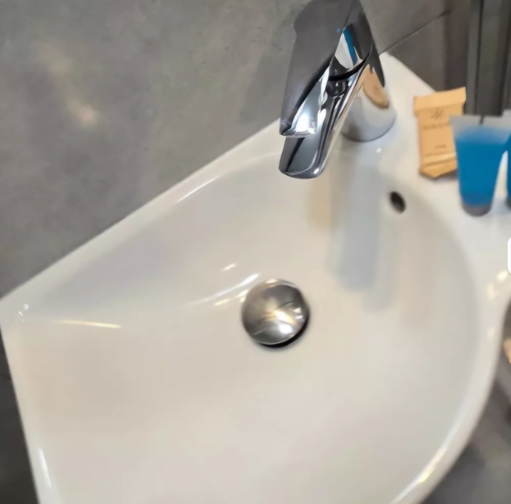 Biała umywalka z chromowaną baterią i zestawem kosmetyków hotelowych na blacie, na tle szarej ściany. Ujęcie z góry, widoczny odpływ umywalki.