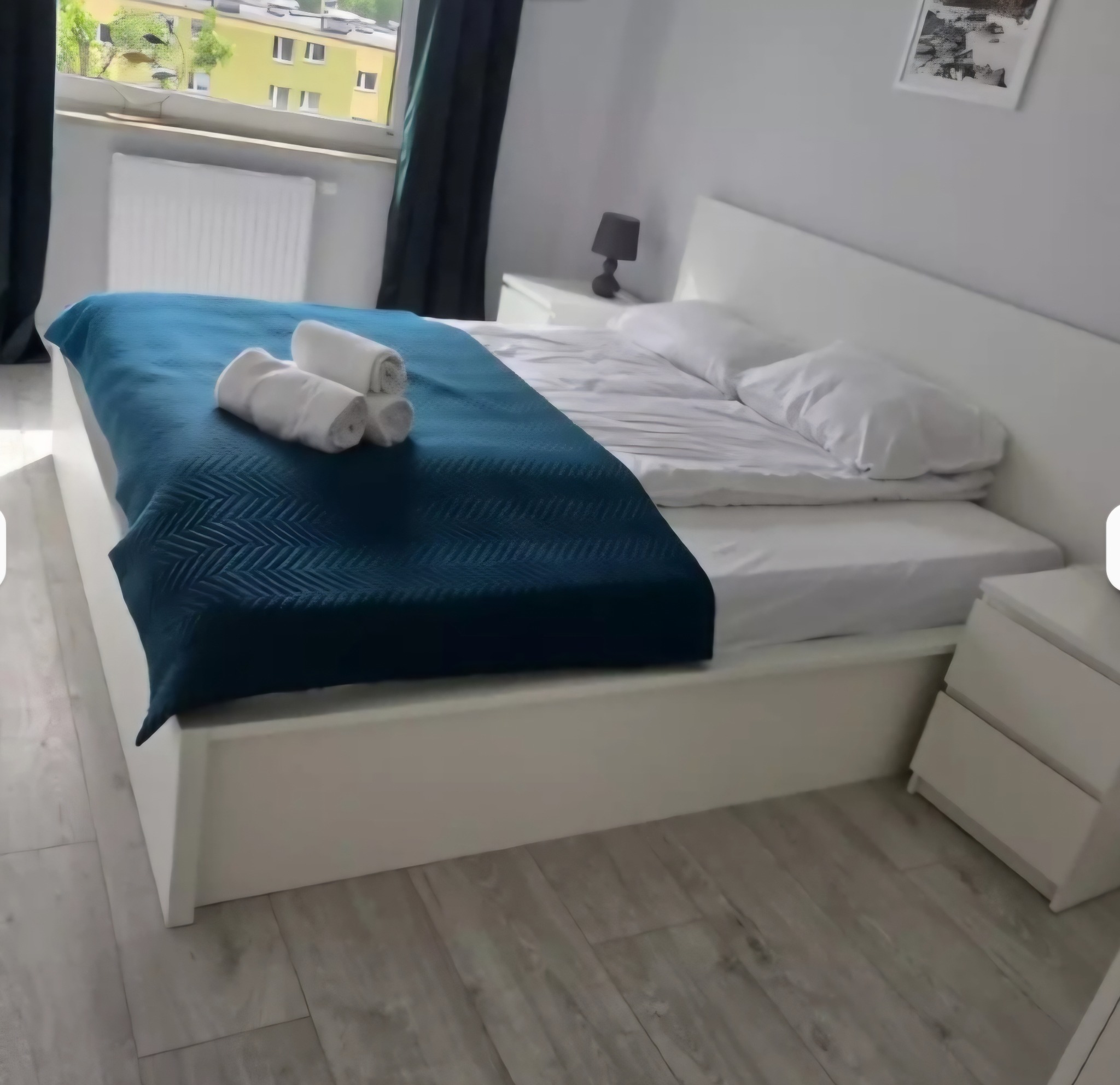 Jasne wnętrze sypialni z białym łóżkiem, turkusową narzutą i zwiniętymi ręcznikami. Widok na okno z zielenią za nim. Minimalistyczny styl, czyste linie i drewniana podłoga.