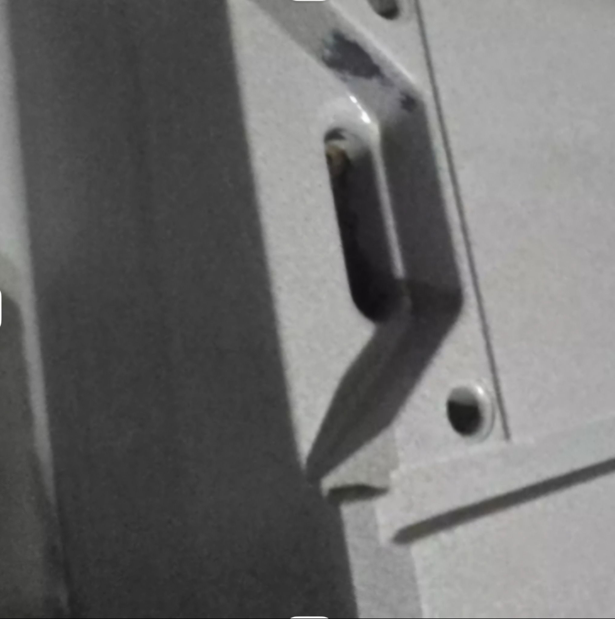 Szary, metalowy element konstrukcyjny z widocznymi otworami i śladami spawania, oświetlony z góry, z fragmentem cienia. Detal z bliska.