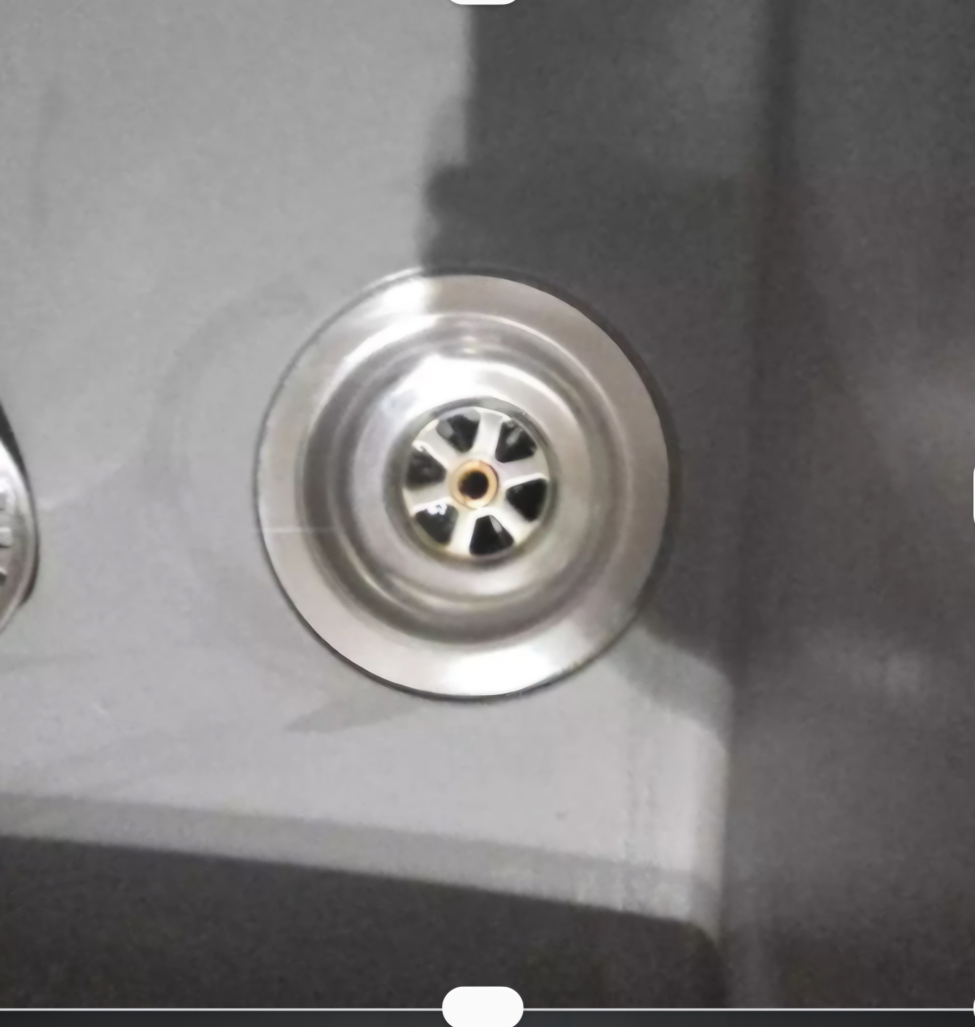 Błyszczący, metalowy odpływ zlewu w zbliżeniu, z widocznymi drobnymi zabrudzeniami wewnątrz. Szary zlew z delikatnym połyskiem, oświetlony z góry.